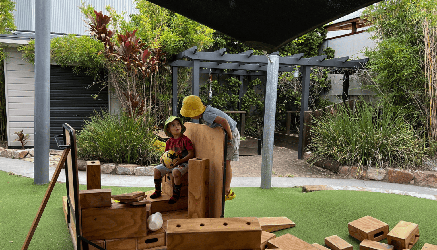 Two young children wearing sun hats are playing with wooden blocks in an outdoor play area. One child is sitting and holding a soccer ball, while the other is climbing on the structure they've built. The scene is set under a shaded canopy with lush greenery and a pergola in the background.