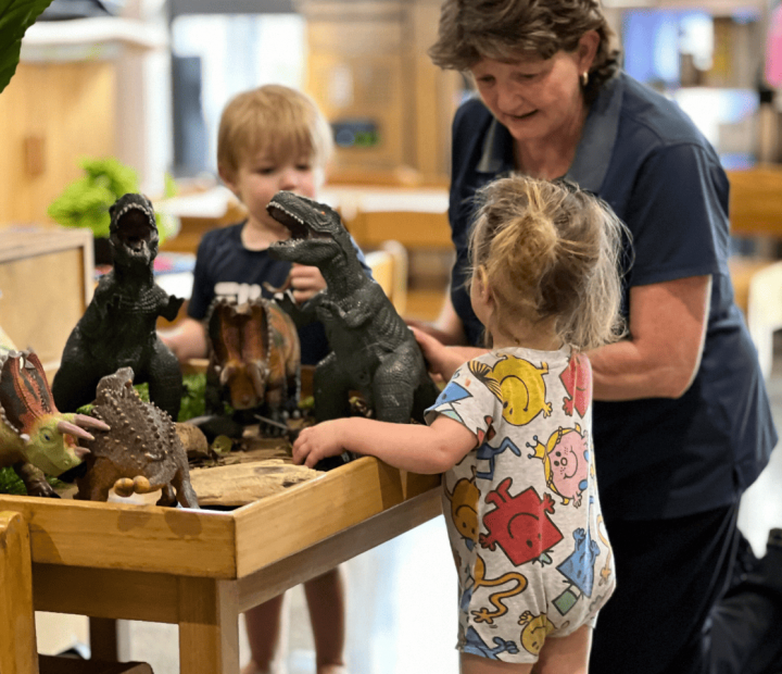 A teacher and two young children are playing with toy dinosaurs on a wooden table. The children, one wearing a colorful onesie, are engaged in imaginative play, while the teacher interacts with them. There are potted plants and various dinosaur figures, including a Triceratops and a Tyrannosaurus Rex, on the table.