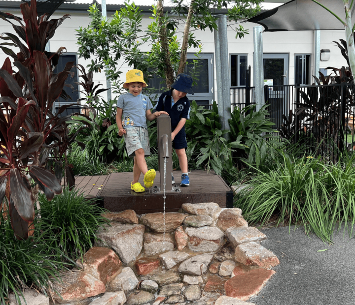 Two young boys are playing at a water pump feature in an outdoor garden area. One boy in a yellow hat is stepping on the pump to release water, while the other boy in a blue hat looks on. The scene is surrounded by lush plants and rocks, with a modern building and shaded play area in the background.